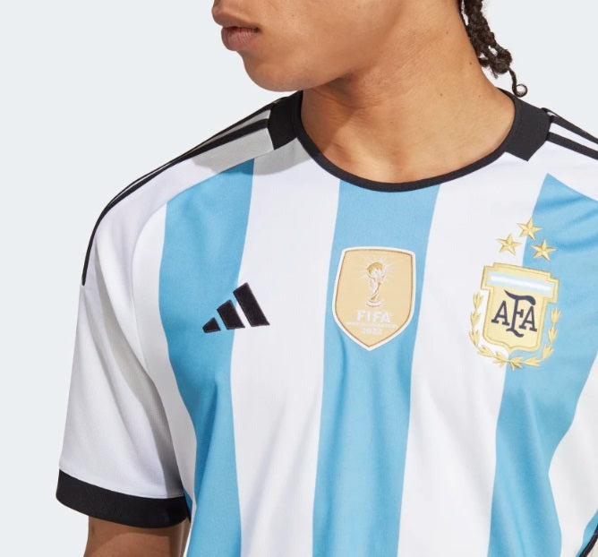 Primer plano del escudo de la Selección Argentina de Fútbol con las tres estrellas doradas bordadas, escudo FIFA World Champions 2018 y adidas logo sobre el tejido oficial del jersey 2022.