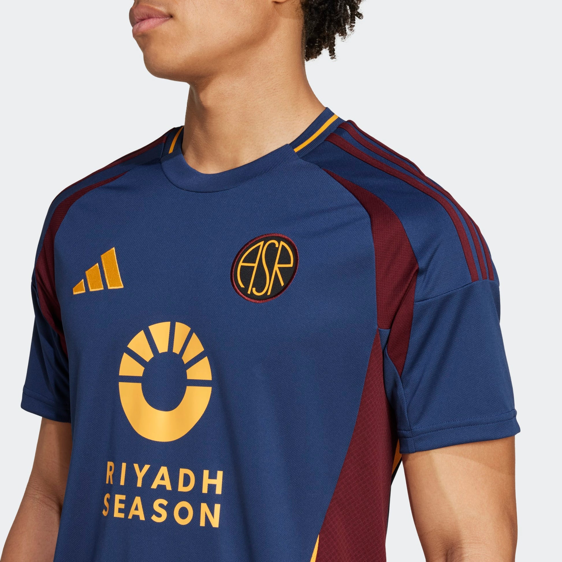 Primer plano del escudo histórico 'ASR' de la AS Roma, bordado con precisión sobre el tejido azul del tercer jersey. A la izquierda, el nuevo logotipo de Adidas en color amarillo vibrante