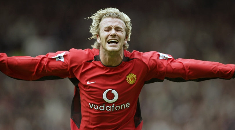 David Beckham celebrando emocionado en la cancha con brazos extendidos luciendo la camiseta roja del Manchester United en Futbol90MX