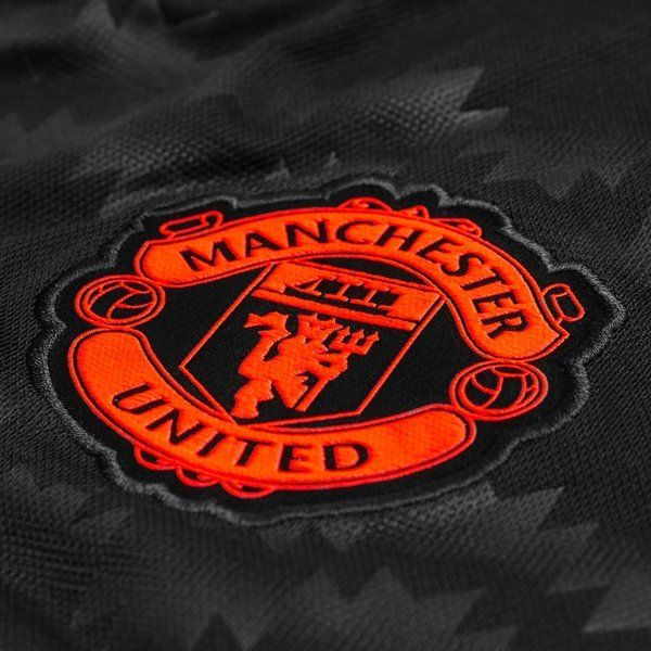 Primer plano macro del escudo oficial del Manchester United bordado en tonos negro y rojo solar sobre la textura de patrón geométrico del tercer kit 2015/16 de Beckham (Ref. AC1445).