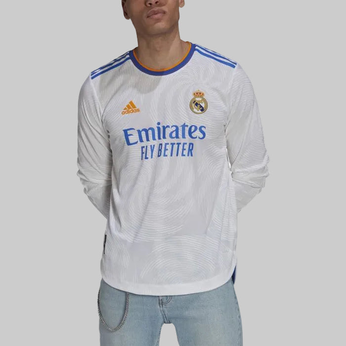 Modelo vistiendo el jersey authentic match de manga larga Adidas del Real Madrid UCL 2021/22 de Modric (Código GR3995) en color blanco con detalles en azul royal y naranja, combinado con jeans, en una presentación exclusiva de Futbol90MX.