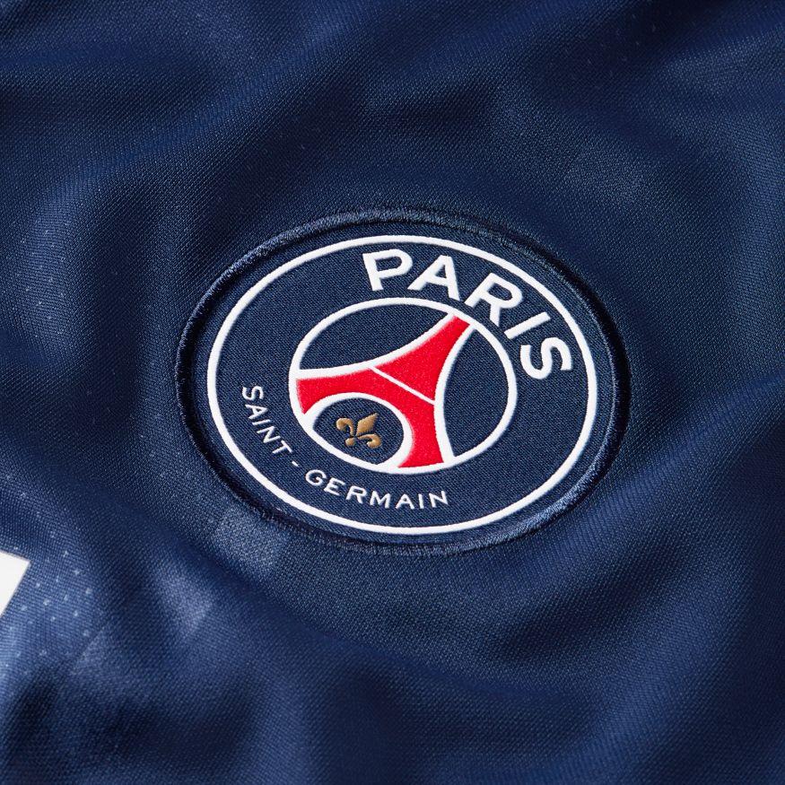 Vista macro de alta precisión sobre el pecho del jersey de local UCL de Mbappe, resaltando el escudo del PSG bordado en el tejido azul marino