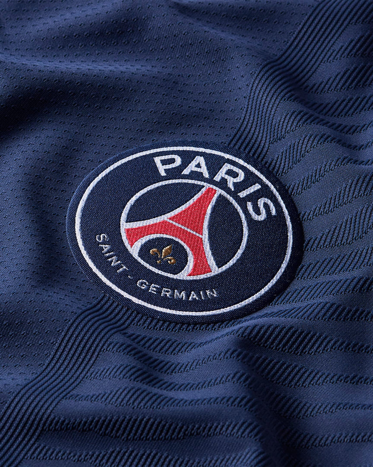 Detalle en primer plano del escudo oficial del Paris Saint-Germain termo aplicado sobre la textura de tejido de rendimiento azul marino del jersey de Mbappe Vapor Match Local UCL 2021/22 . Se aprecian los hilos dorados y rojos del emblema sobre la base azul.