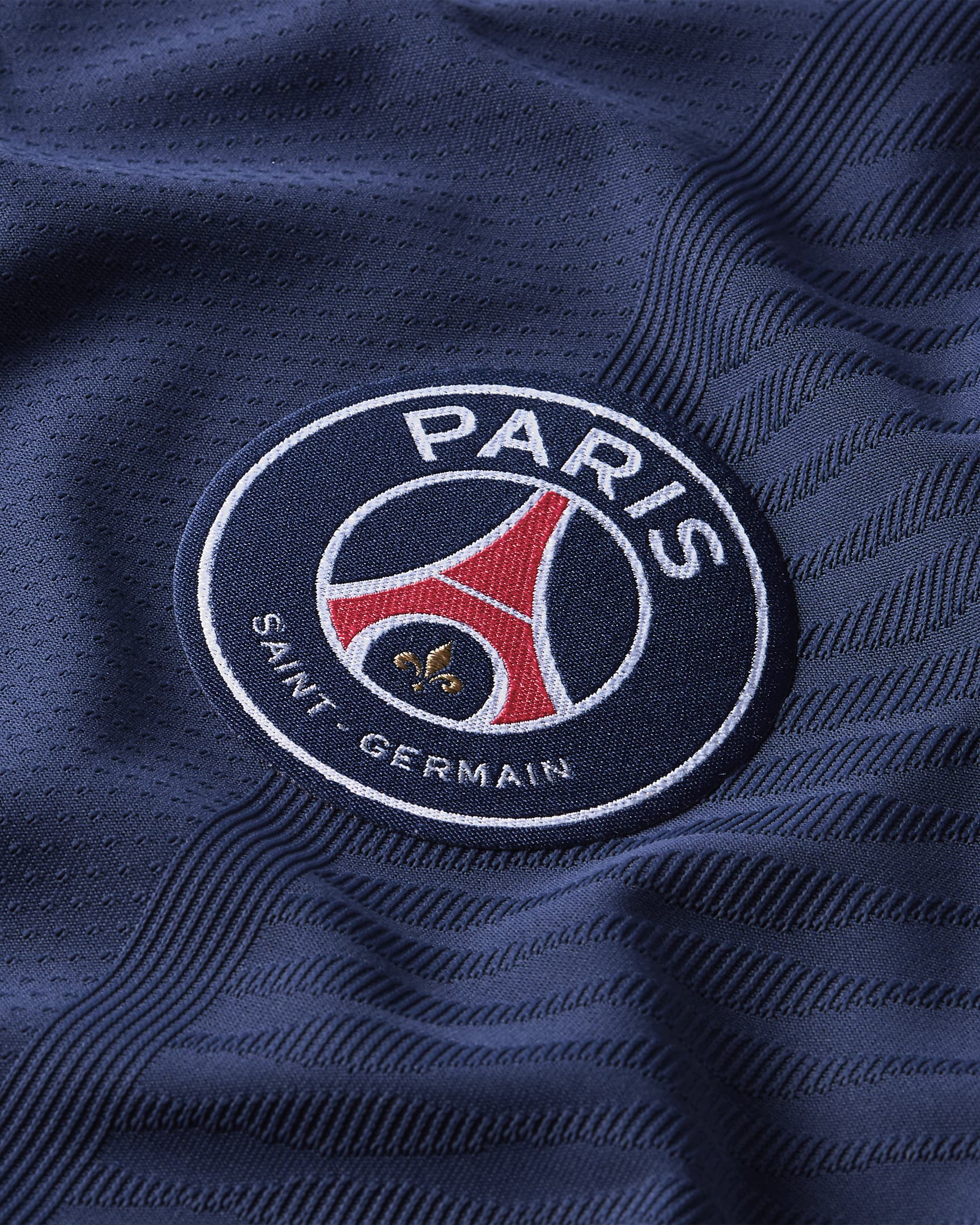 Detalle en primer plano del escudo oficial del Paris Saint-Germain termo aplicado sobre la textura de tejido de rendimiento azul marino del jersey de Mbappe Vapor Match Local UCL 2021/22 . Se aprecian los hilos dorados y rojos del emblema sobre la base azul.
