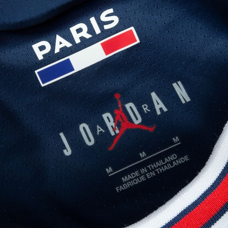 Macrofotografía de la etiqueta del cuello interior del jersey Vapor Match Jordan Mbappe PSG Local UCL 2021/22. Se distingue el logotipo Jumpman rojo sobre el texto "JORDAN", la bandera francesa, y detalles de la talla y origen de fabricación en Tailandia