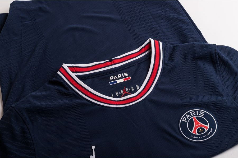 Vista frontal en ángulo del jersey Jordan Mbappe PSG Home UCL 2021/22 Authentic. Muestra el cuello redondo de punto elástico azul con franjas tricolores rojas y blancas, y la parte superior del escudo del club termo aplicado.