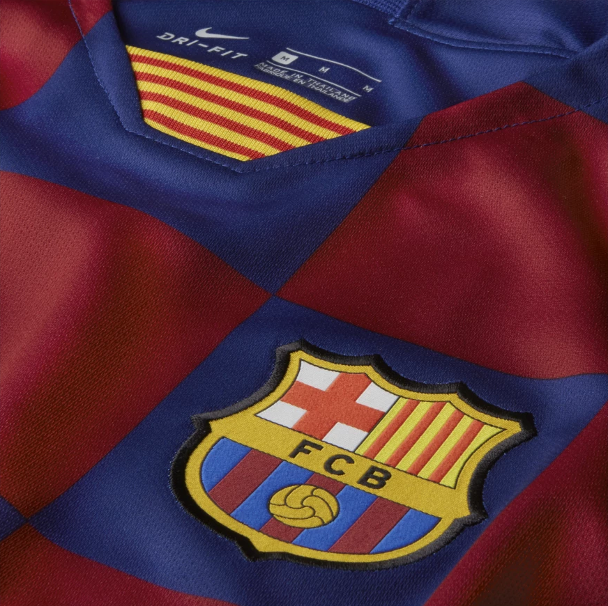 Primer plano de alta definición del jersey local de Iniesta del FC Barcelona 2019/20, destacando el escudo bordado del club sobre el patrón de cuadros azulgrana y el detalle del cuello en V con la bandera de la Senyera y el logotipo Nike Dri-FIT en Futbol90MX.