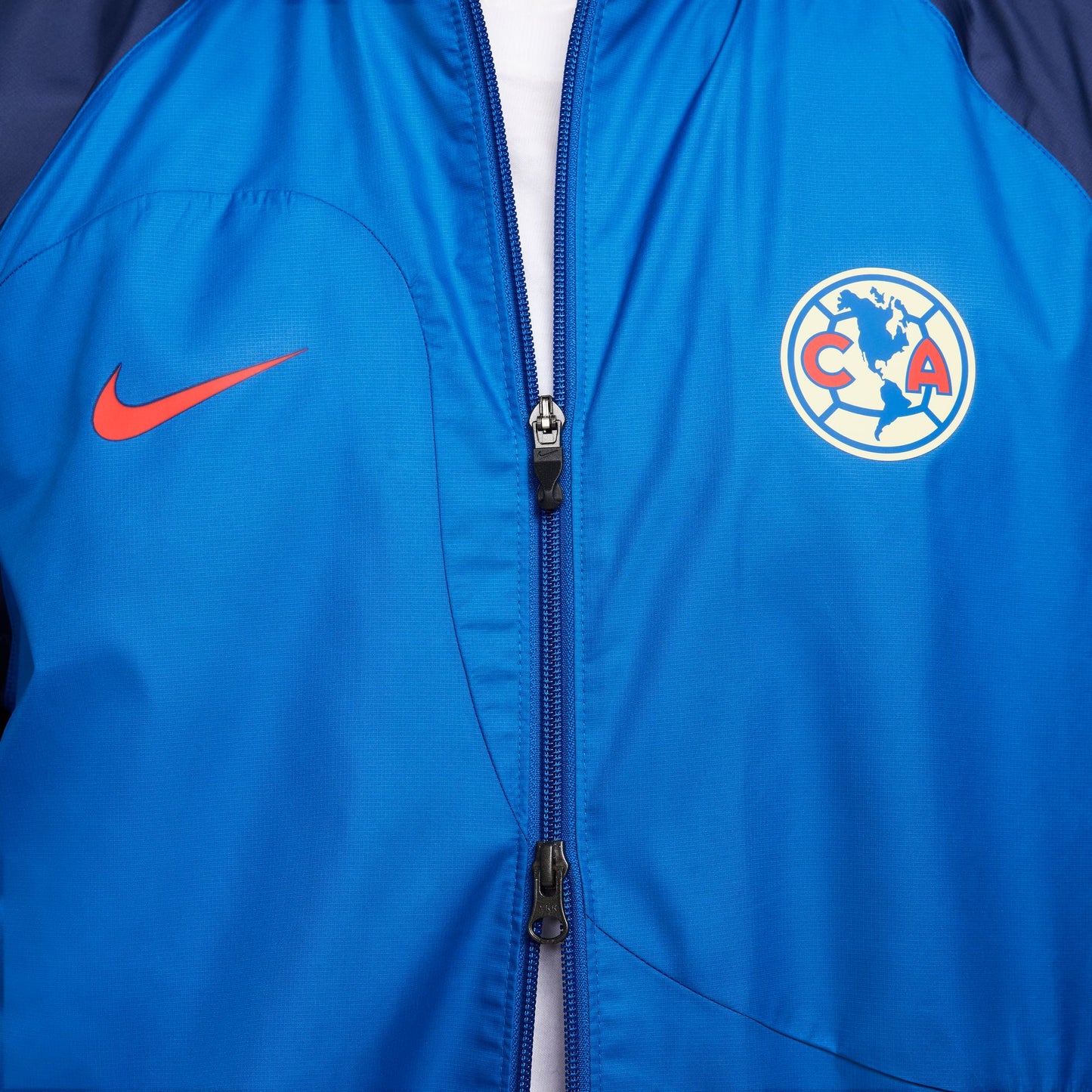 Acercamiento al escudo del Club América y el Swoosh de Nike en color carmesí aplicado con precisión técnica sobre tejido Woven repelente al agua.