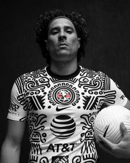 Retrato en blanco y negro de Guillermo Ochoa vistiendo el jersey de arte azteca 2020/21 del Club América, sosteniendo un balón de fútbol clásico y mirando a cámara con la identificación de Futbol90MX