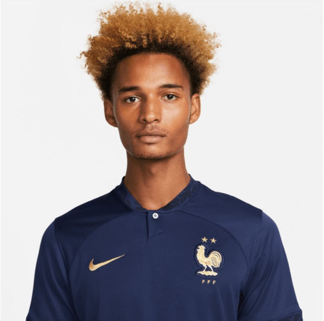 Primer plano del escudo de la Federación Francesa de Fútbol y el logo de Nike en acabado dorado metálico sobre el tejido azul marino del Jersey de Local Mundial 2022 de Mbappe