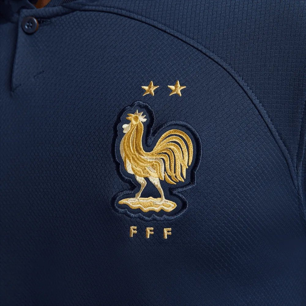 Primer plano del escudo de la Federación Francesa de Fútbol en acabado dorado metálico sobre el tejido azul marino del Jersey de casa manga larga de Mbappe mundial 2022