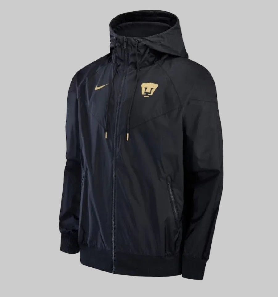 Vista tres cuartos en ángulo frontal de la chamarra Nike Windrunner de Pumas UNAM 2023, destacando el icónico panel frontal chevron a 26 grados, codigo DX7963-014 stock Futbol90MX