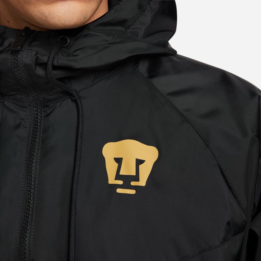 Acercamiento al pecho de la rompevientos Nike de Pumas 2023, mostrando el puma estilizado de la UNAM termoaplicado en oro.