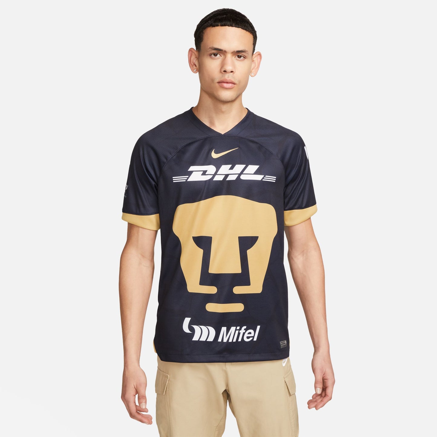 Vista frontal del jersey Pumas UNAM 2023-24 versión de visita. Diseño en azul obsidiana con patrón de piedra apilada y logotipos de patrocinadores DHL y Mifel en color blanco nítido.