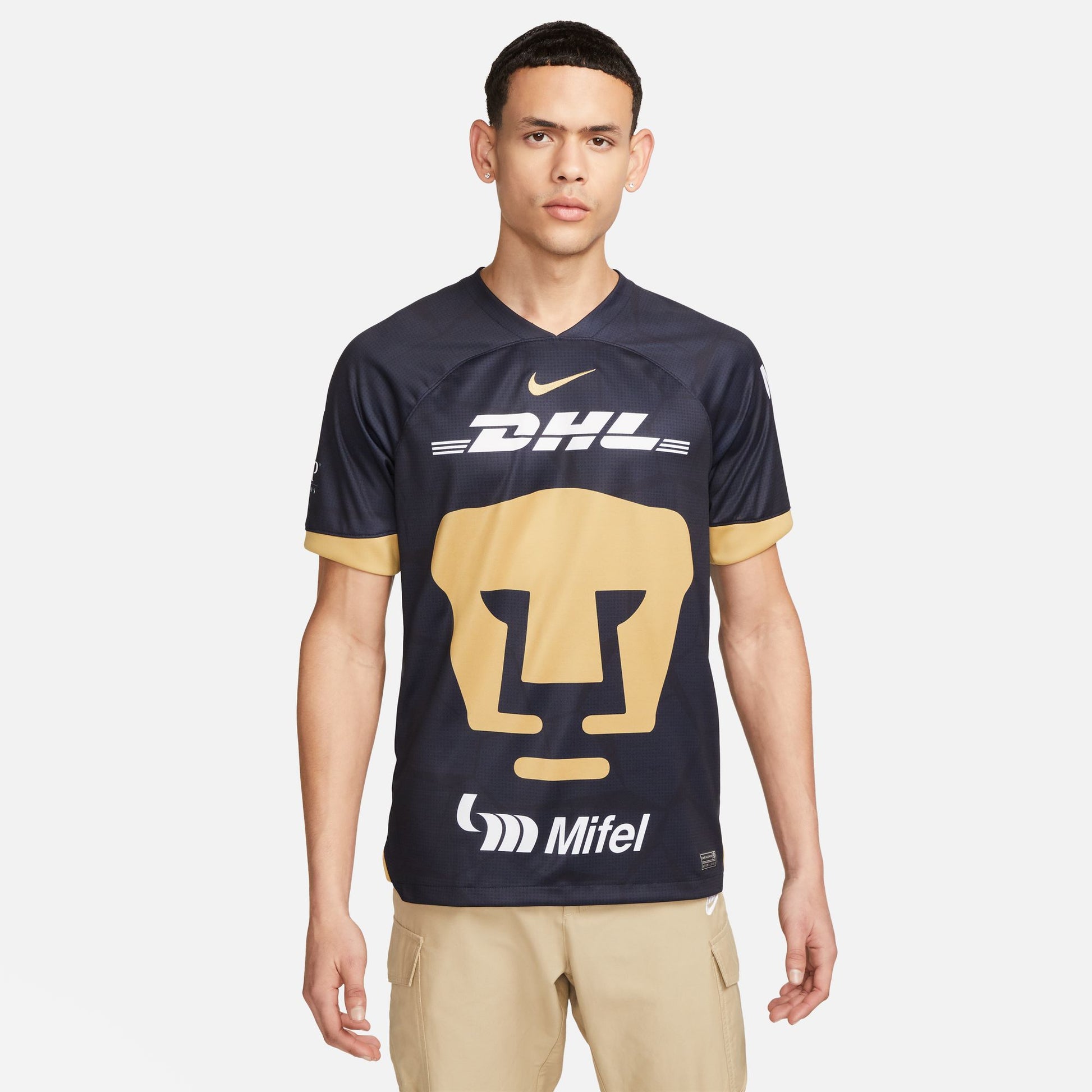 Vista frontal del jersey Pumas UNAM 2023-24 versión de visita. Diseño en azul obsidiana con patrón de piedra apilada y logotipos de patrocinadores DHL y Mifel en color blanco nítido.