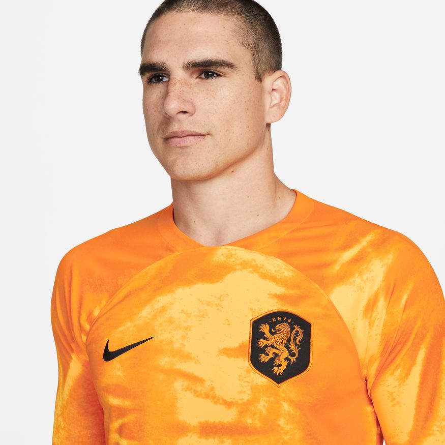 Plano detalle del escudo de la Federación Neerlandesa (KNVB) y el logo Nike Swoosh sobre el tejido técnico naranja, exhibiendo la construcción premium del activo.