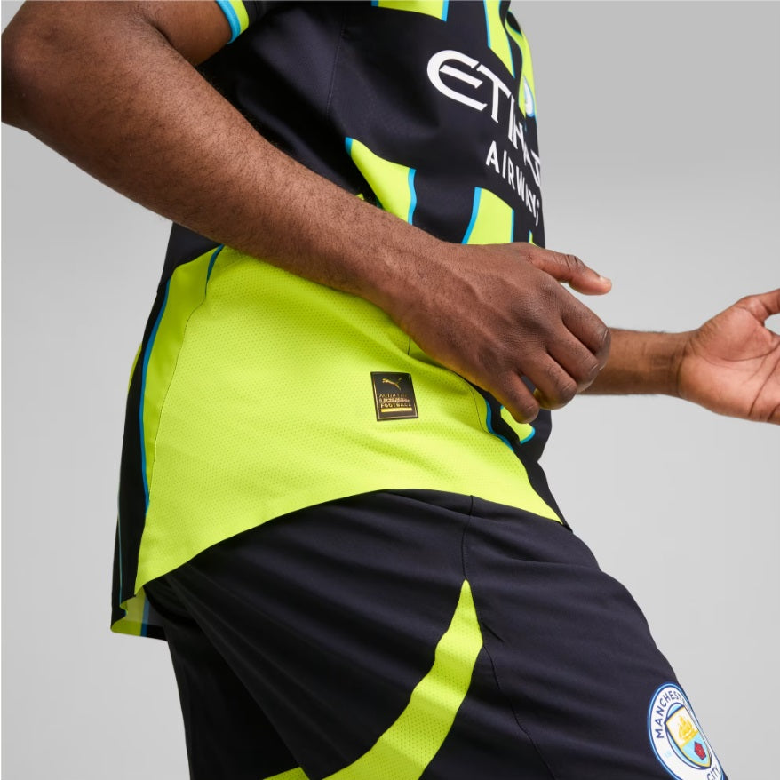 Plano detalle lateral en movimiento mostrando el parche dorado de autenticidad PUMA Authentic Licensed Football en el dobladillo inferior del jersey Manchester City Authentic Match Away UCL 2024/25 de Haaland (Ref. 775085 02) en Futbol90MX