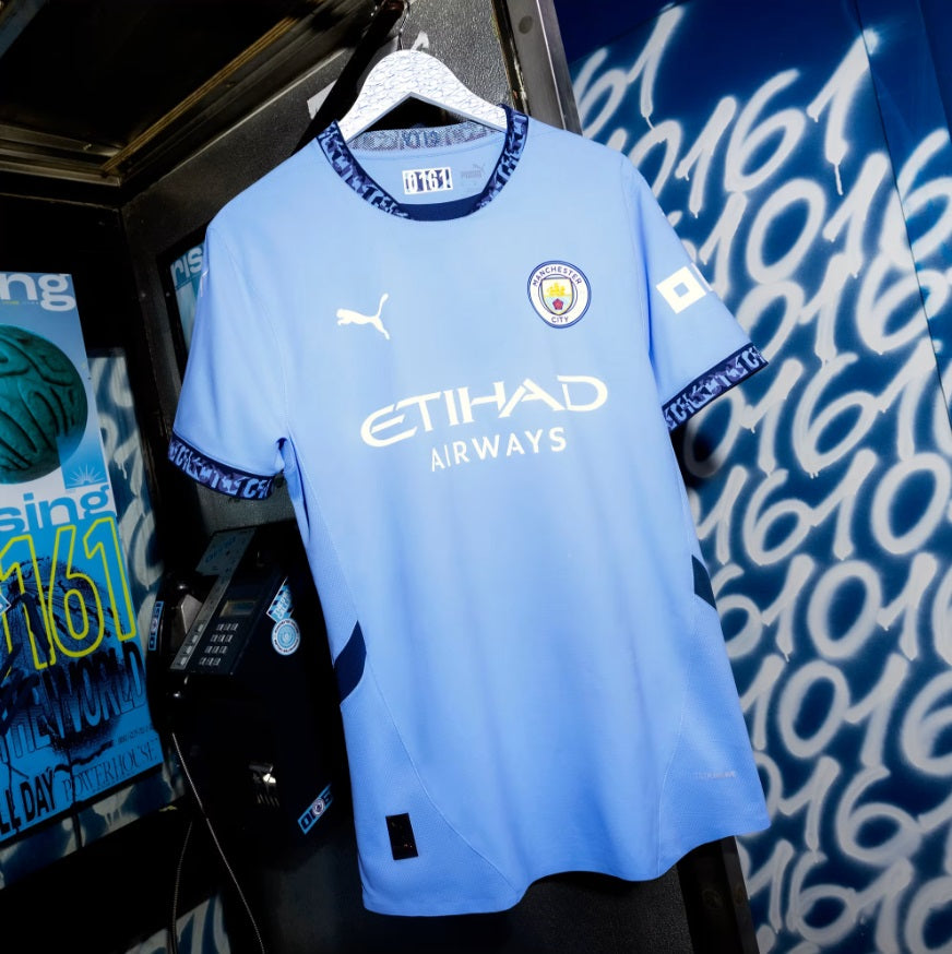 Camiseta Puma Erling Haaland Manchester City Home 2024/25 (Ref. 775075 01) en color City Blue colgada en perchero contra un fondo urbano con inscripciones "0161" en Futbol90MX