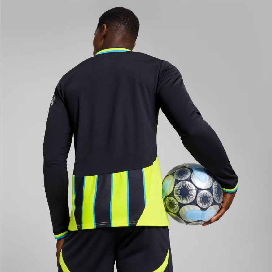 Vista posterior de un modelo sosteniendo un balón, mostrando el diseño trasero del jersey away de manga larga del Manchester City 2024/25, en Futbol90MX