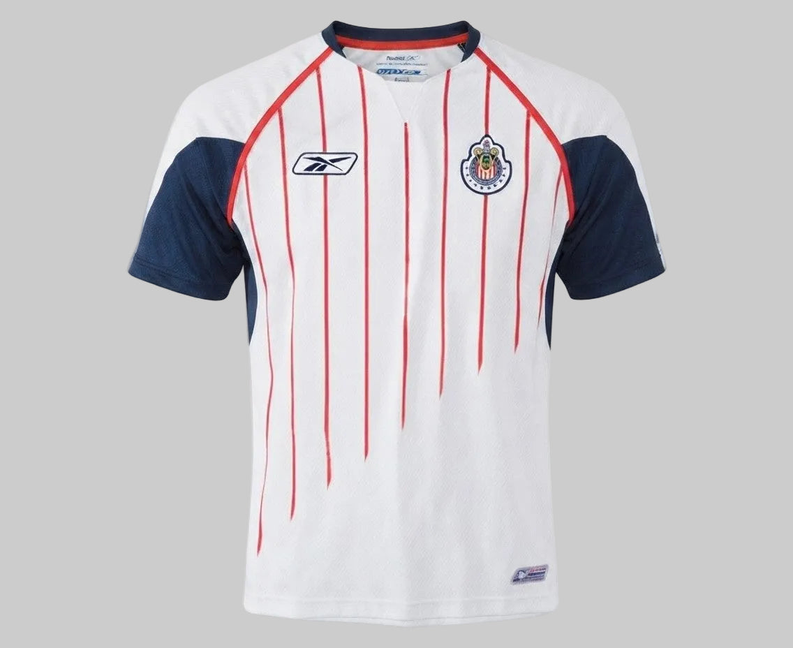Primer plano del pecho del jersey de visita de Chivas 2005/06, destacando la textura de la tela blanca, el escudo oficial con estrellas y el logotipo de Reebok, stock Futbol90MX